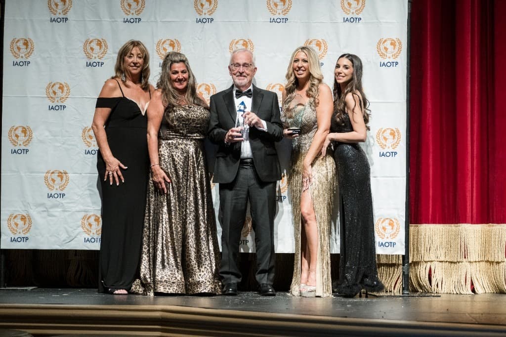 Prof. Richard Larson in a tuxedo holding a crystal award on the IAOTP gala stage with four women in formal gowns